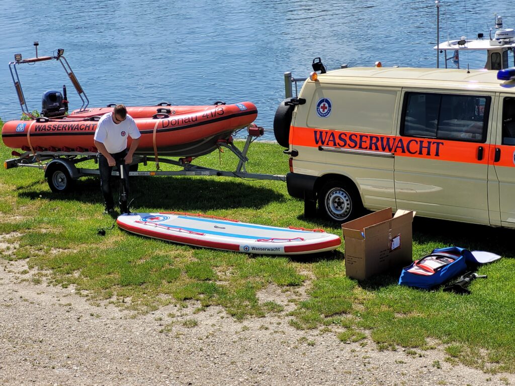 Wasserwachtbus mit Schlauchboot. Im Vordergrund ein Mitglied der Wasserwacht, der ein SUP aufpumpt.